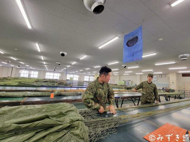 整備工場の点検台で落下傘を畳む整備中隊隊員と、その後ろで状況を解説するために控えている説明係の隊員